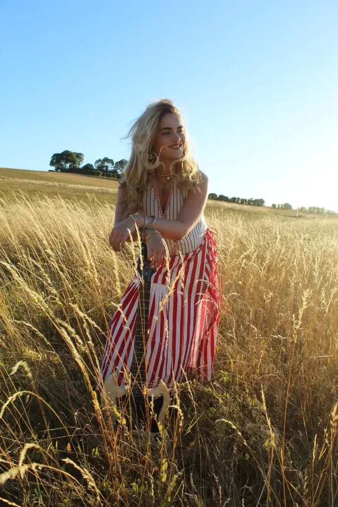 Indiana leaning on a guitar in tall summer grass, laughing, in a red and white striped dress.