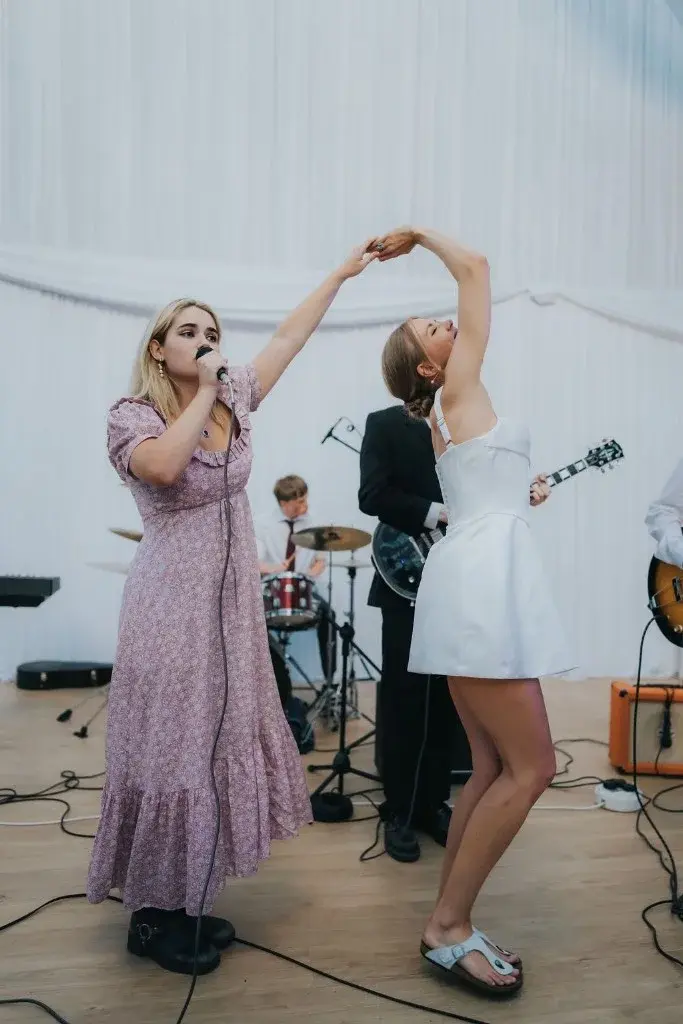 Indiana singing on stage in a pink floral dress, holding hands with a dancing friend, the band behind them.